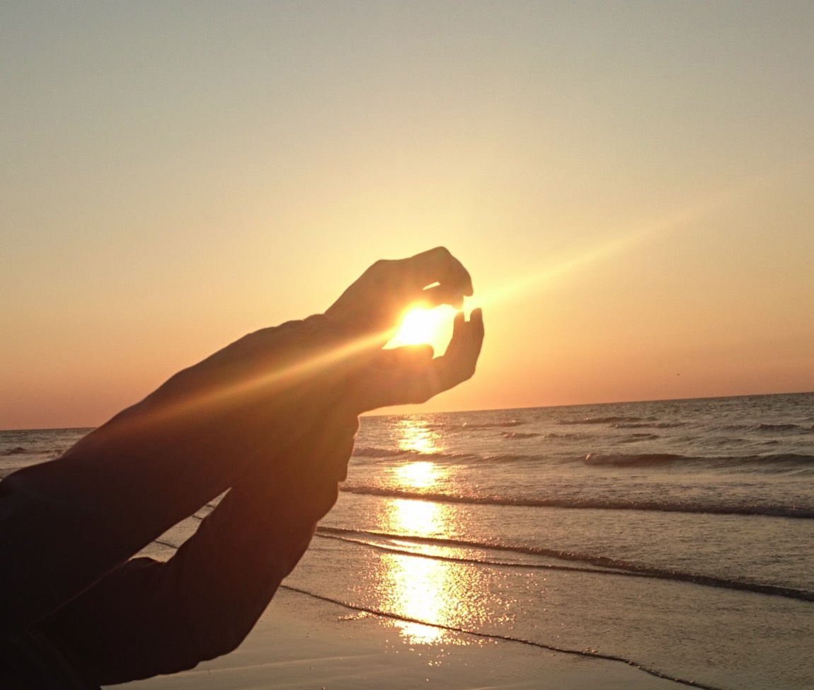 Auf dem Bild sieht man zusammen gelegte Hände, durch die die Sonne über dem Meer leuchtet.