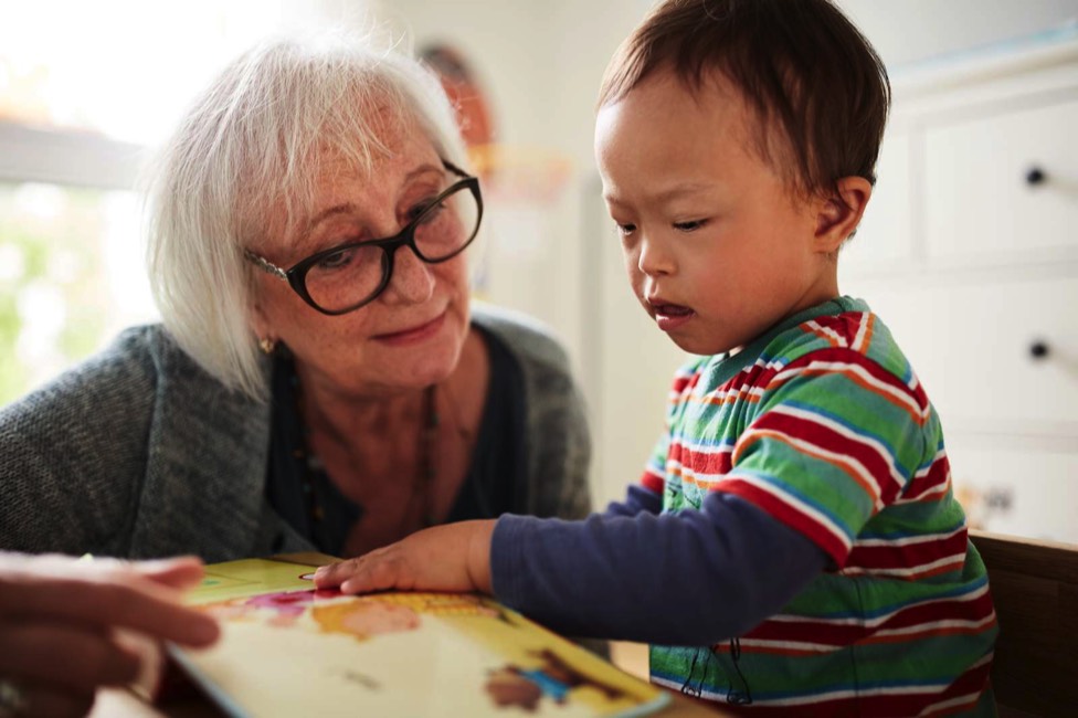 Auf dem 9. Bild sieht man eine ältere freiwillige Frau, die sich zusammen mit einem kleinen Jungen ein Bilderbuch anschaut.