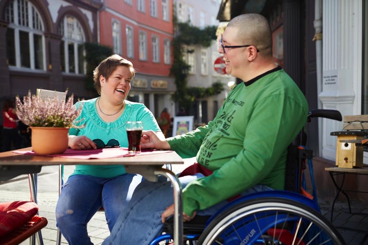Auf dem 3. Bild sieht man eine Freiwillige und einen Mann im Rollstuhl draußen in einem Café.